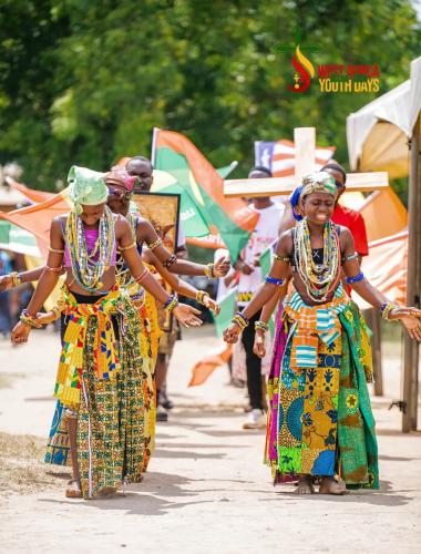 Zwei junge Frauen in farbenfroher traditioneller Kleidung tanzen bei den „West Africa Youth Days Accra 2026“, während Teilnehmende mit Flaggen und einem Holzkreuz hinter ihnen gehen.
